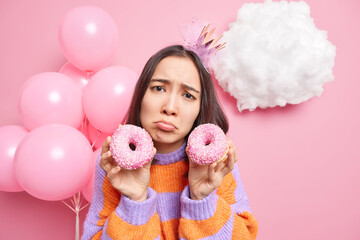 Frustrated displeased young Asian woman frowns face purses lips looks with gloomy expression holds two glazed donuts poses at party with bunch of balloons in background. Unhealthy food concept