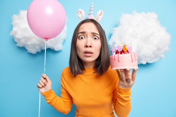 Wall Mural - Worried brunette Asian woman looks nervously at camera dressed in casual orange turtleneck holds delicious cake and inflated balloon isolated over blue background with clouds above. Festive occasion