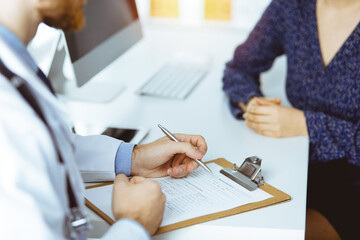 Unknown bearded doctor and patient woman discussing current health examination while sitting in sunny clinic, close-up