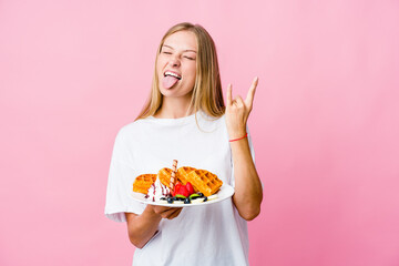 Wall Mural - Young russian woman eating a waffle isolated showing rock gesture with fingers