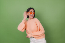 Young Stylish Woman In Casual Peach Sweater And Orange Glasses Isolated On Green Olive Background Curious Try To Hear What You Saying With Hand By Ear Copy Space