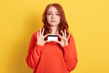 Close Up Portrait Of Upset Frowning Girl Holding Credit Card With Both Hands, Showing It To Camera, Feeling Perplexed, Standing Over Yellow Background, Being Sad, Has Not Enough Money For Shopping.
