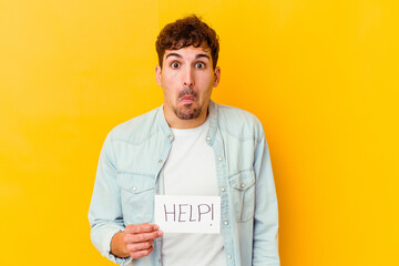 Wall Mural - Young caucasian man holding a help placard isolated shrugs shoulders and open eyes confused.