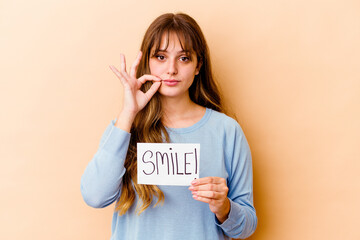 Wall Mural - Young caucasian woman holding a Smile placard isolated with fingers on lips keeping a secret.