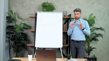 Man business coach trainer standing near whiteboard flip chart tells talk speak teaches company manager employees student remote online webcam video conference call chat laptop computer in home office