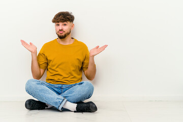 Young Moroccan man sitting on the floor isolated on white background confused and doubtful shrugging shoulders to hold a copy space.