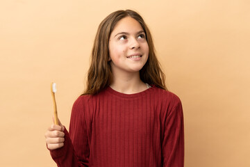 Little caucasian girl brushing her teeth isolated on beige background thinking an idea while looking up