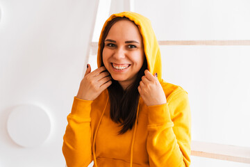 Wall Mural - Portrait of young woman posing on white background. Pretty brunette in yellow hoodie near stepladder on background of white wall.