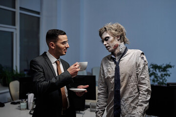 Wall Mural - Young elegant businessman offering cup of coffee to zombie colleague in office environment