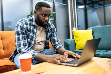 Wall Mural - Serious african american freelancer is using laptop, working on important project. Concentrated male employee sitting in modern office, developing new app or business plan for company