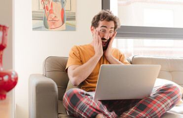 young bearded man feeling happy, excited and surprised, looking to the side with both hands on face and sitting with a laptop