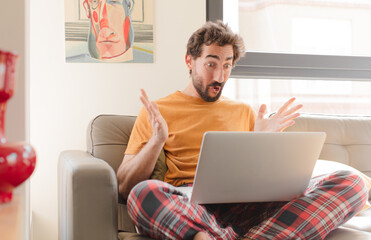 young bearded man feeling extremely shocked and surprised, anxious and panicking, with a stressed and horrified look and sitting with a laptop