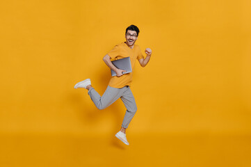 Full length photo of a cheerful funny caucasian smart young man in eyeglasses, stylishly dressed, holding laptop, smiling and jumping high running on isolated orange background