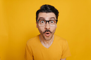 Portrait of amazed foolish cheerful caucasian guy in glasses and in an orange t-shirt, cheerfully looking at the camera with a distorted face, standing against an isolated orange background