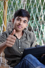 Wall Mural - Indian Handsome boy with Tablet resting in hammock outdoors and showing  Thumbs up	

