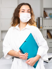 Young elegant businesswoman in face mask standing in white office and holding a folder