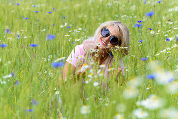 Beautiful middle-aged female blonde in sunglasses in a field among flowers, daisies and cornflowers. Portrait of a happy smiling woman enjoying summer. 