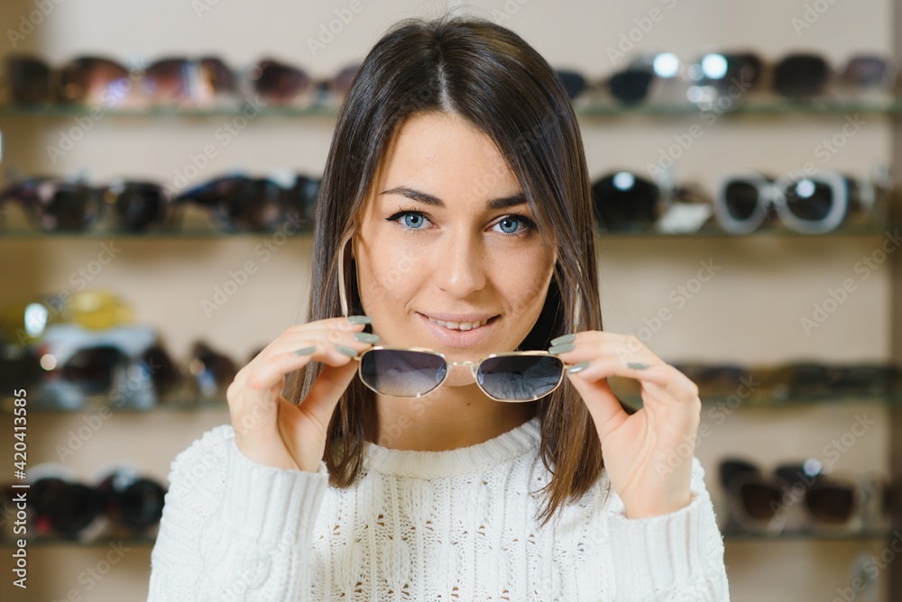 Which one is better fit me. Portrait of cheerful hesitating woman in optician store, making decision, holding stylish sunglasses, choosing what she should buy. Great discounts for buying two pairs - obrazy, fototapety, plakaty 