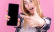 © Studio Nova - Surprised happy blonde woman in shirt showing blank smartphone screen and pointing at it while looking at camera with open mouth on pink background