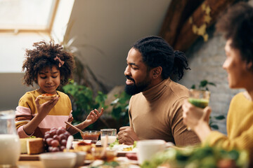 Happy African American father talking to daughter during breakfast at dining table.