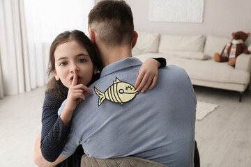 Cute little girl sticking paper fish to father's back at home