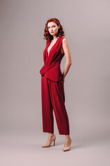 Wall Mural - Feminist look idea. Sexy ginger woman in bright red jumpsuit with deep neckline. Women’s clothing, fashion concept. Burgundy evening gown. Sensual portrait. Natural looking makeup and wavy hair.