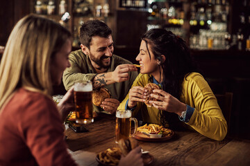 happy woman having fun while boyfriend is feeding her with french fries during lunch in a pub.