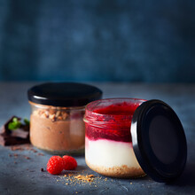 Two Homemade Easy Cheesecakes In Jars With Chocolate Mousse And Raspberry Curd Sauce On A Dark Blue Background. Healthy Dessert. Selective Focus. Homemade Raw Vegan Pudding.