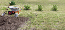 Patch Of Grass And Dug Up Against The Background Of Watering Can, Shovel, Rake And Garden Wheelbarrow Against The Backdrop Of A Garden And A Vegetable Garden. Break After Processing The Site