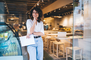 Happy female manager with menu in hand standing in doorway of local cafeteria and smiling, pretty Caucaisan small owner enjoying business lifestyle for working with franchise takeaway industry