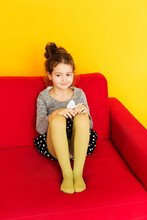 A little sweet girl with dark hair in a gray sweater and small skirt is sitting on a red sofa on a yellow background. Girl smiling