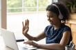 © fizkes - Ready to start. Positive biracial teen female in headset sit by laptop waving hand to camera greet teacher tutor at distant video lesson. Young black woman student learning online via web conference