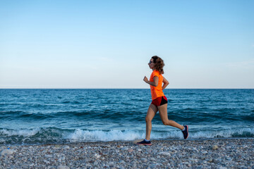 A slender woman in a bright orange T-shirt runs along the beach.