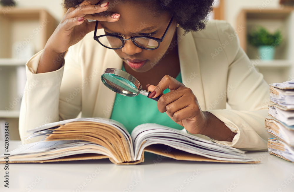 Closeup portrait of concentrated young african american woman auditor with magnifying glass scrutinizing financial documents at desk with paper folder pile in office. Investigation and audit checkup - obrazy, fototapety, plakaty 