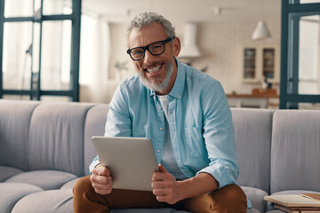 Happy senior man using digital tablet and looking at camera while sitting on the sofa at home