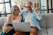 © gstockstudio - Modern senior couple in casual clothing smiling and using laptop while bonding together at home