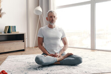 Senior Man In Sport Clothing Meditating While Sitting On The Floor In The Lotus Position At Home