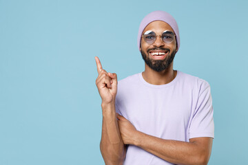 Wall Mural - Young smiling unshaven black dark-skinned african american man 20s in violet t-shirt hat glasses point index finger aside on copy space area mock up isolated on pastel blue background studio portrait.