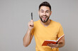 © ViDi Studio - Young caucasian dreamful bearded insighted active happy student smart man 20s wearing casual yellow basic t-shirt writing down in exercise book diary isolated on grey color background studio portrait.