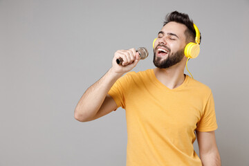 Wall Mural - Young caucasian smiling bearded handsome student happy man 20s wearing casual yellow basic t-shirt listen to music in headphones sing song in microphone isolated on grey background studio portrait.