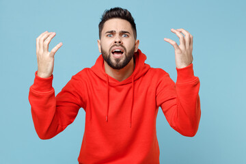 Wall Mural - Young caucasian angry indignant nervous irritated stressed bearded man 20s wearing casual red orange hoodie spreading hands isolated on blue color background studio portrait People lifestyle concept.