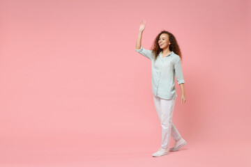 Wall Mural - Fulll length young black african american happy smiling friendly curly woman 20s in blue shirt looking camera walk going waving hand greet someone isolated on pastel pink background studio portrait.