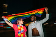 © MyMicrostock/Stocksy - Happy multiracial couple raising LGBT pride flag in street at ni