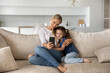 © fizkes - Smiling young Caucasian mother and little 7s daughter relax on sofa at home talk on video call on smartphone. Happy mom and small girl child use cellphone make self-portrait picture together.