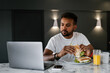 © Alina Hvostikova/Stocksy - Ethnic man eating sandwiches and watching video on laptop
