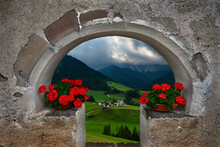 Green Meadows, Village And Mountains Through Old Wall  Arch With Red Flowers. Ancient Stone Wall In Stormy Weather. 