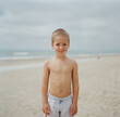 © Jakob Lagerstedt/Stocksy - Cute young boy standing on a beach in his swim trunks