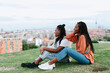 © Jimena Roquero/Stocksy - Two young women sitting on a hill looking at a city landscape