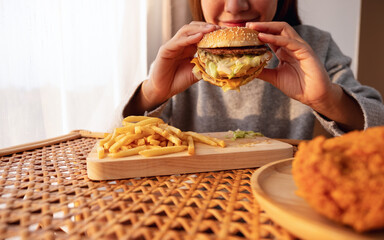 Wall Mural - Closeup image of a woman holding and eating hamburger and french fries with fried chicken on the table at home