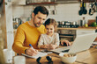 © Drazen - Working father taking notes while daughter is sitting on his lap at home.